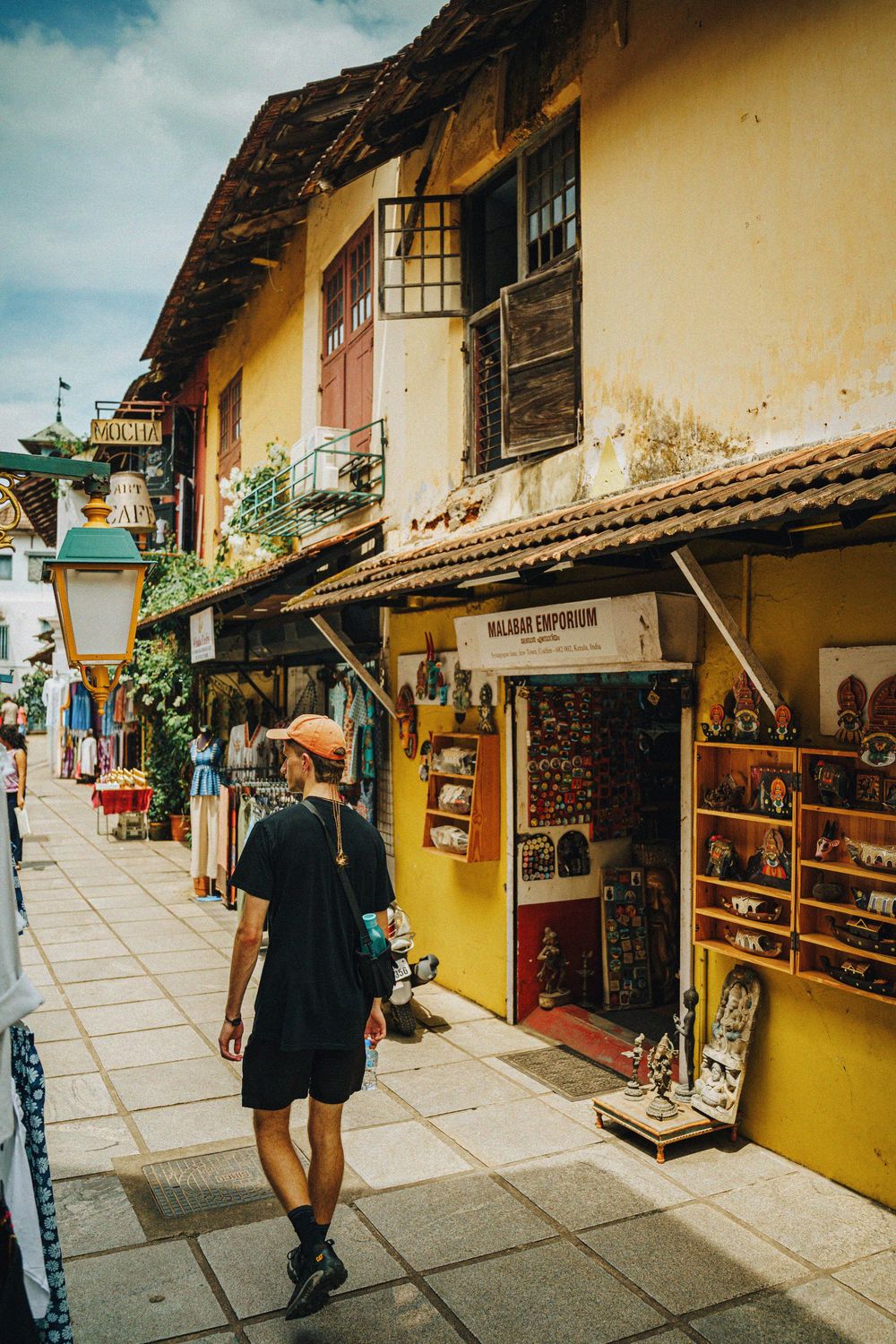 Tourist walking past colorful Malabar Emporium souvenir shop on a narrow street in Fort Kochi, India.
