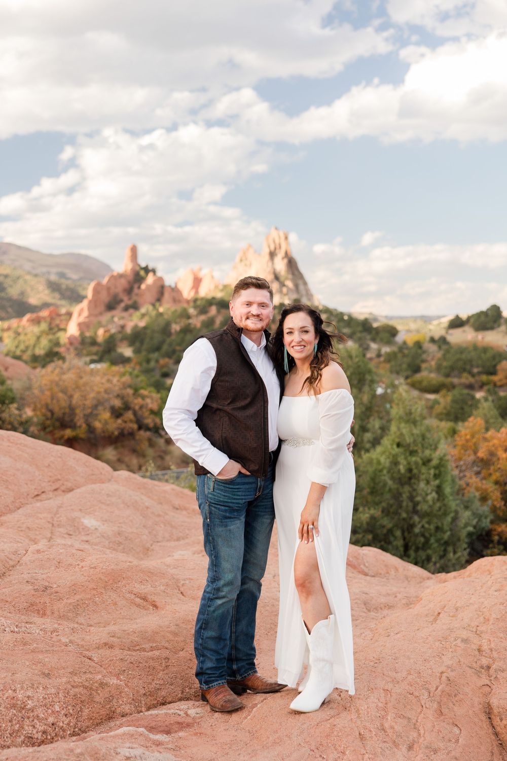 A couple poses together on red rocks at Garden of the Gods park during sunset with mountain views in the background.