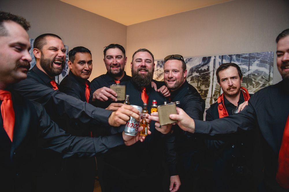 Groomsmen in black suits with orange ties toast together with shot glasses at a wedding celebration.
