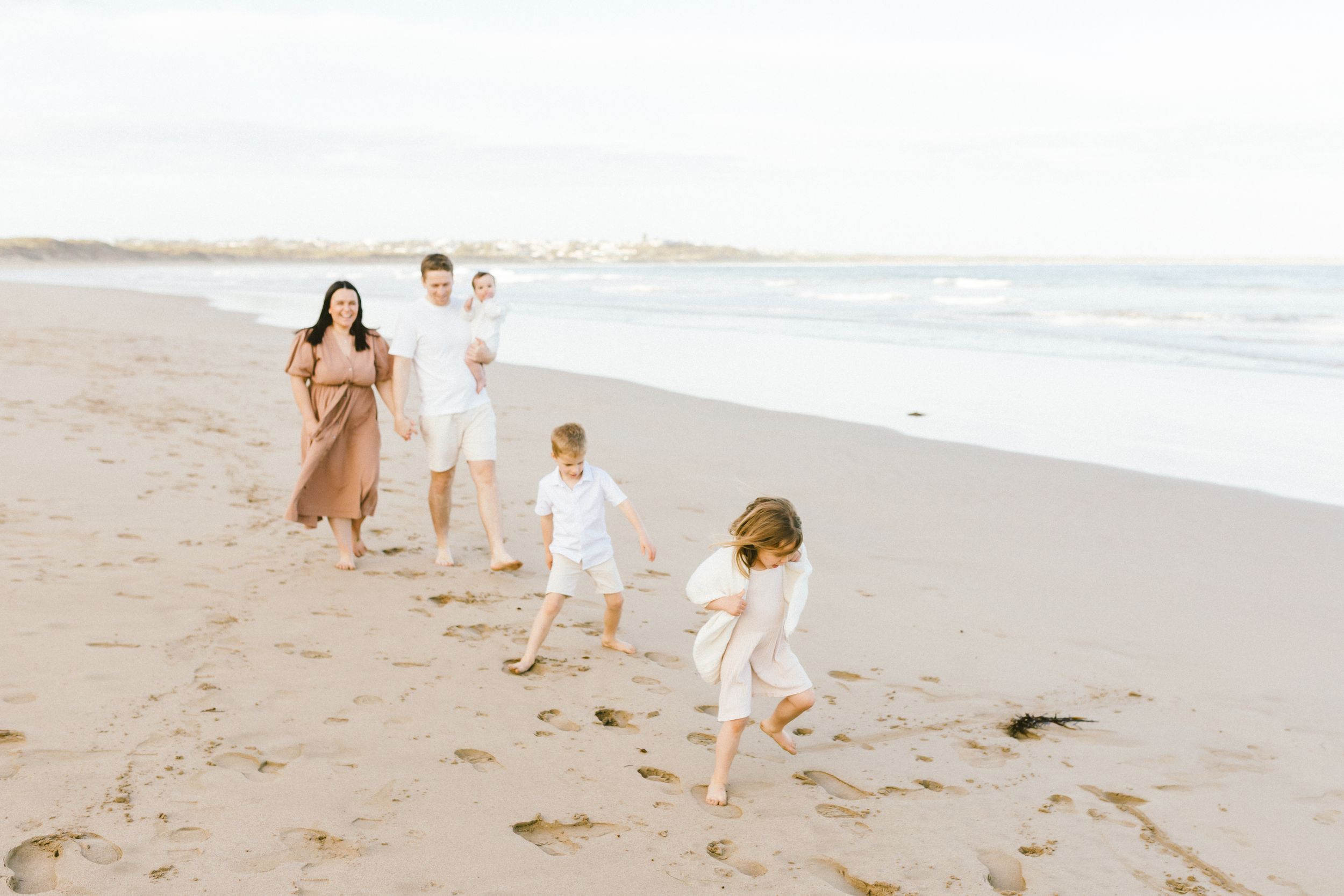 Family wearing white outfits walking together along a sunny beach shoreline during golden hour.