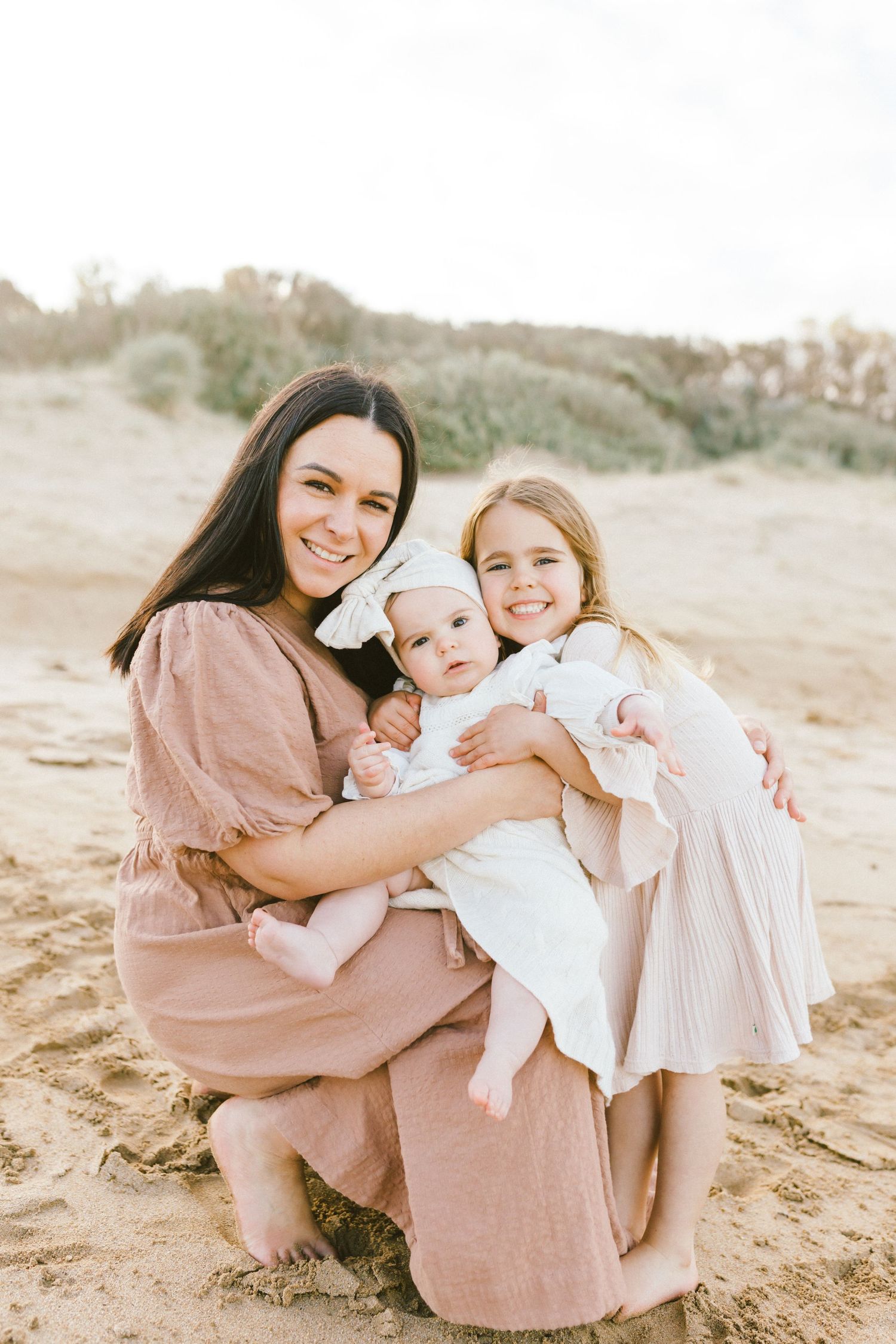 A family in soft neutral tones sits together on sandy ground during an outdoor photo session at sunset.