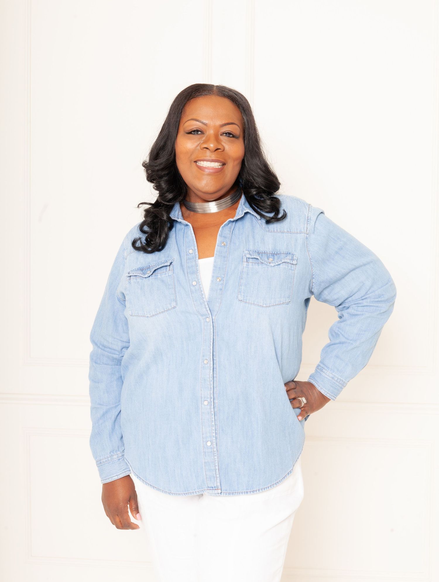 Casual professional portrait in light denim shirt and white pants against white background.