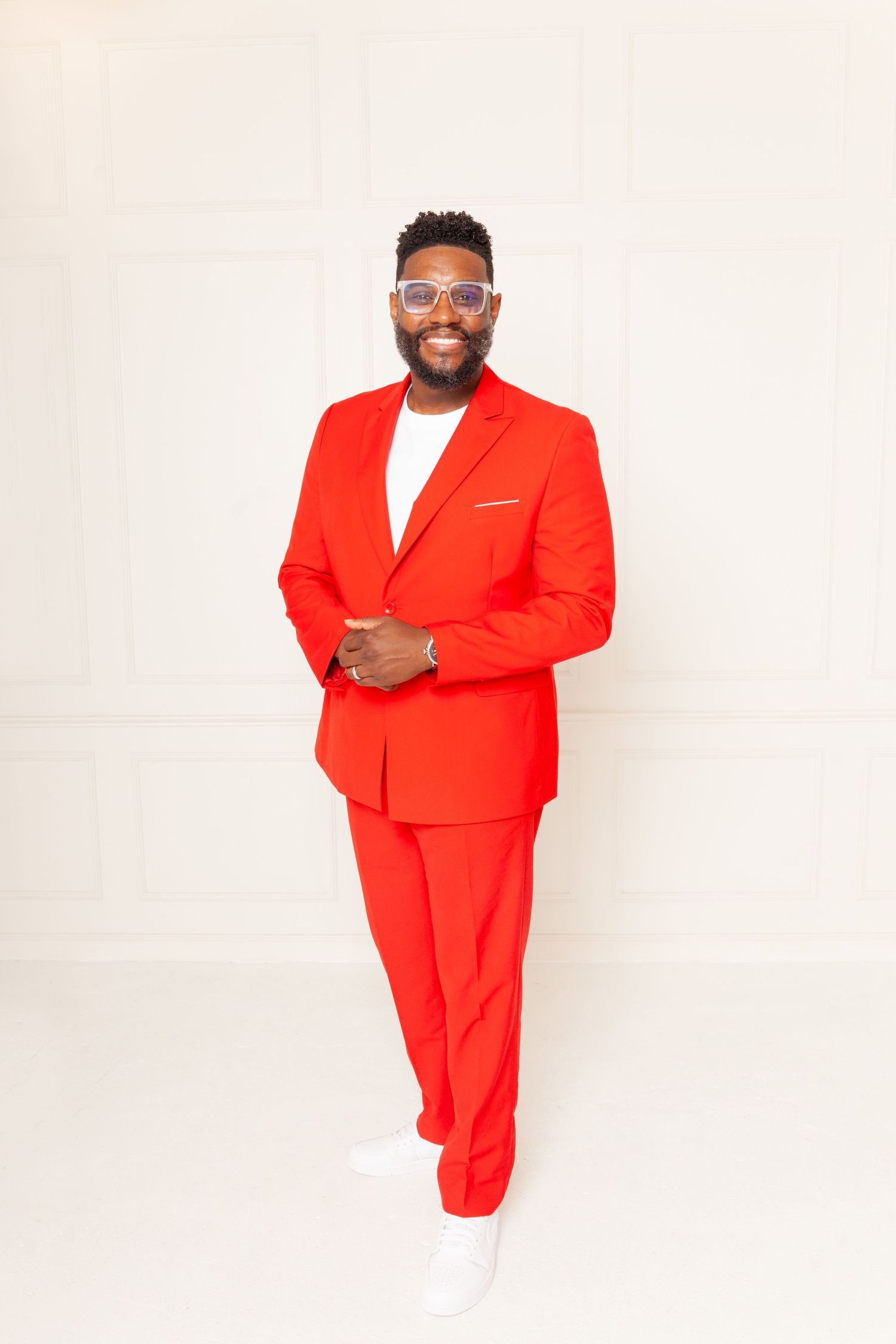 Confident pose in bright red business suit with white shirt and sneakers against white backdrop.