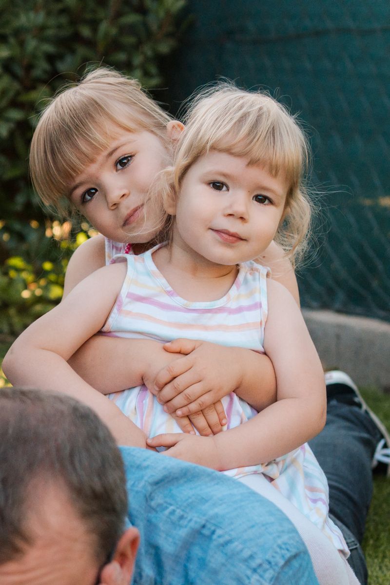Young sisters in a pink-white sleeveless dress hugging in their Rouse Hill home's outdoor setting with greenery during a family photoshoot