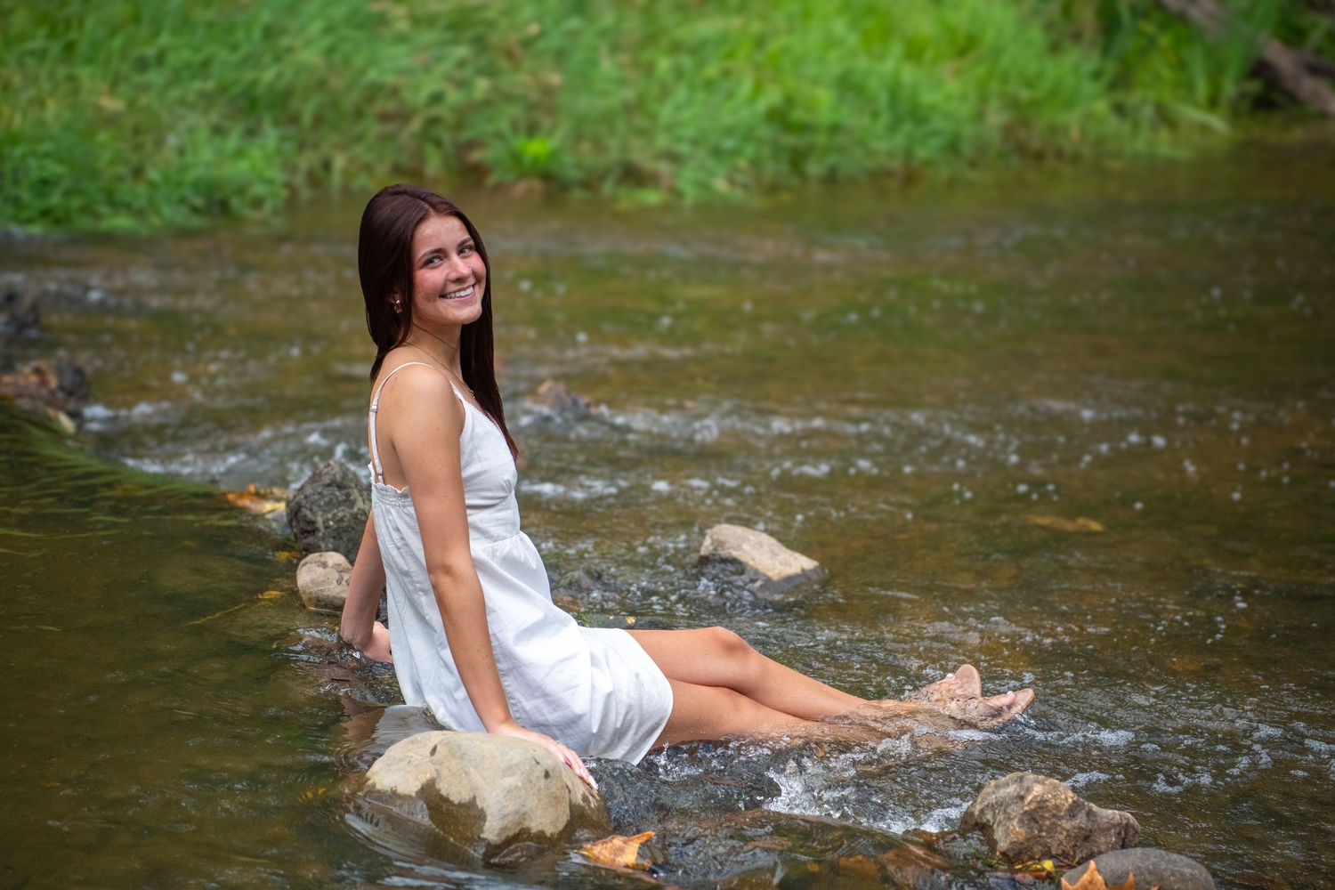 Woman in white dress sitting in flowing stream water on sunny summer day creating splashing movements.
