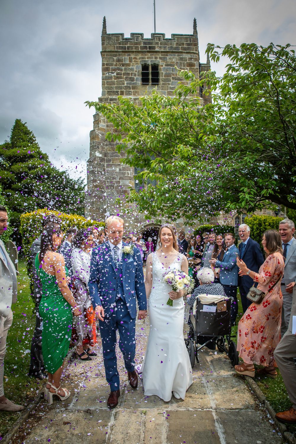 Bride and groom walk through confetti shower outside historic stone church surrounded by cheering wedding guests.