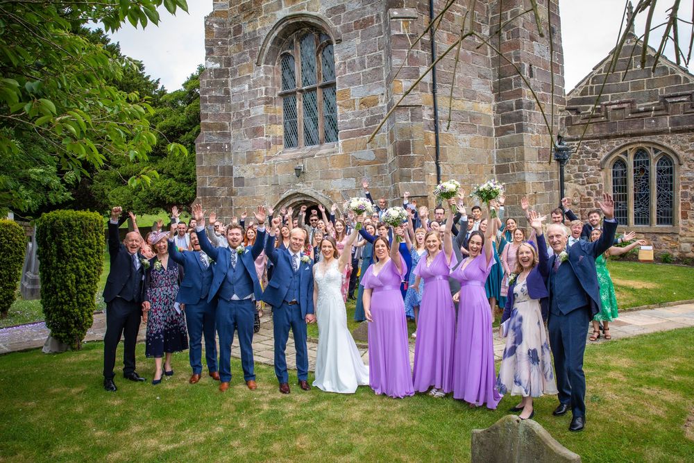 Wedding party celebrates outside stone church with bridesmaids in purple dresses raising hands joyfully.