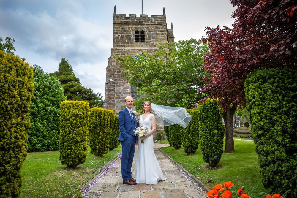 Bride and groom pose on garden path before historic stone church tower with manicured hedges.
