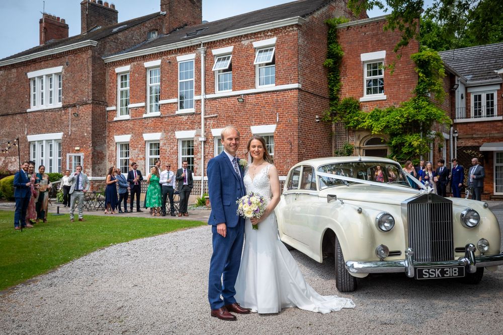 Bride and groom stand beside vintage white Rolls Royce outside elegant red brick manor house.