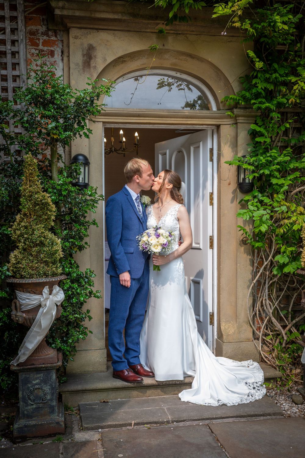 Bride and groom share a kiss in arched doorway of ivy-covered stone manor house entrance.
