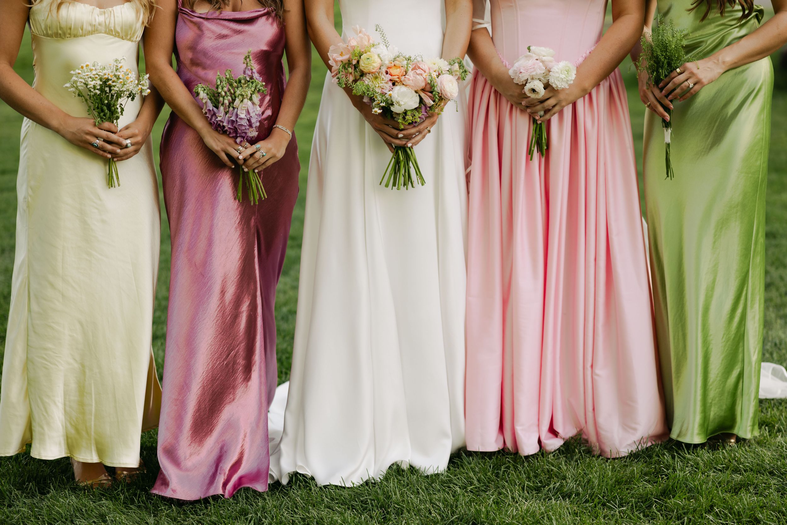 A row of bridesmaids in pink and green dresses holding floral bouquets at a wedding ceremony.