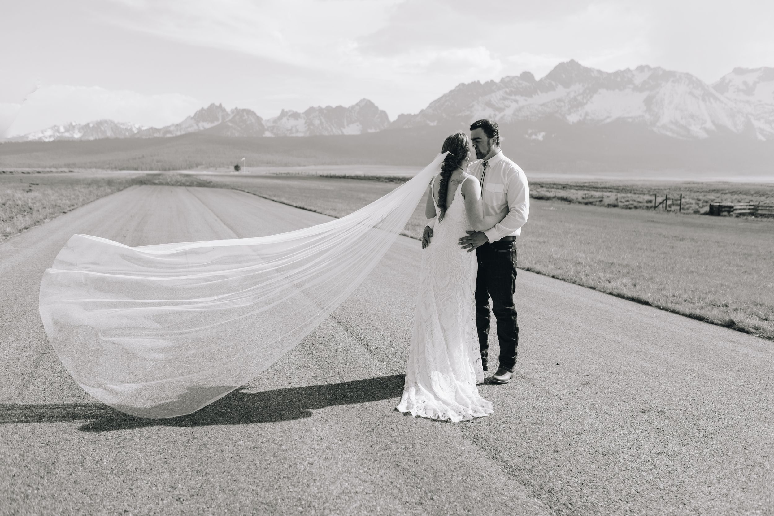A couple in wedding attire stands on a road with mountains in the background as a long veil flows in the wind.