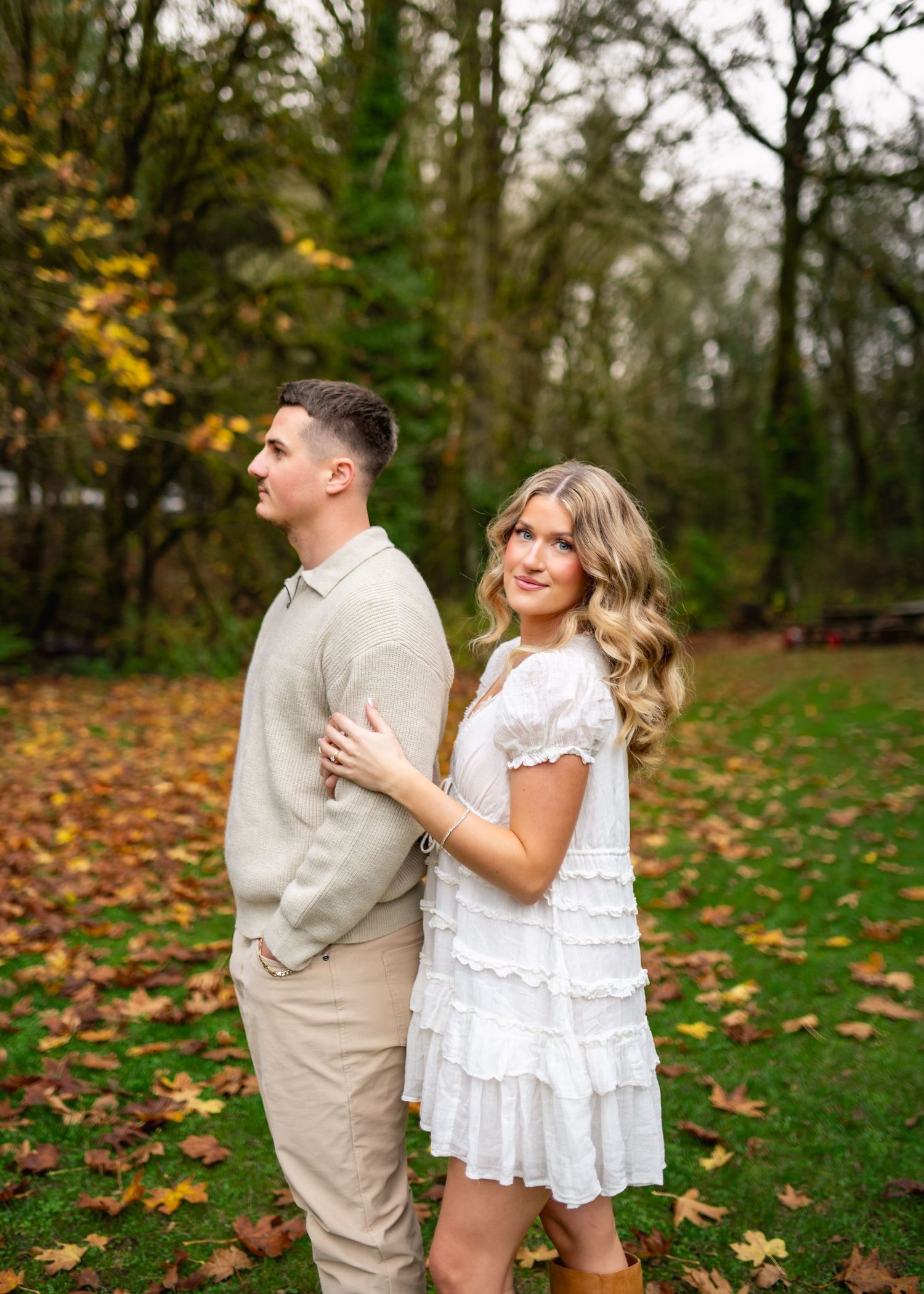 A couple in casual autumn attire shares a romantic moment among fallen leaves in a wooded outdoor setting.