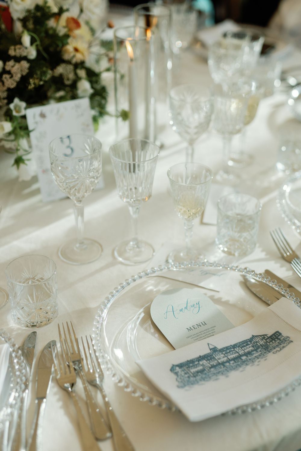 Elegant white table settings with crystal glassware and delicate place cards create a refined wedding reception atmosphere.