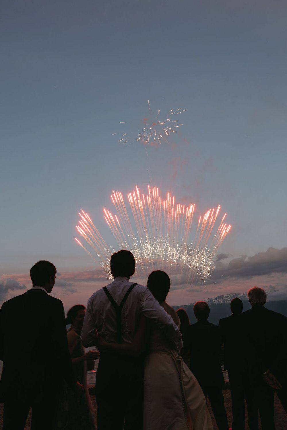 Silhouetted crowd watches colorful fireworks display bursting in twilight sky during outdoor celebration.