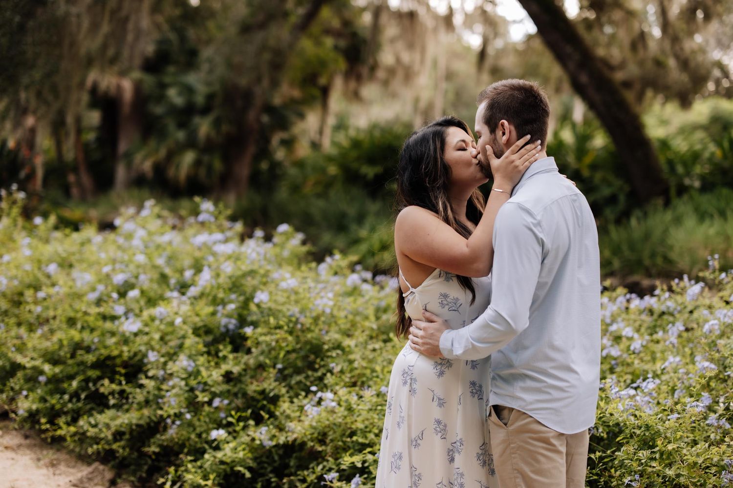 A romantic couple embraces in a lush garden setting surrounded by white wildflowers and Spanish moss.
