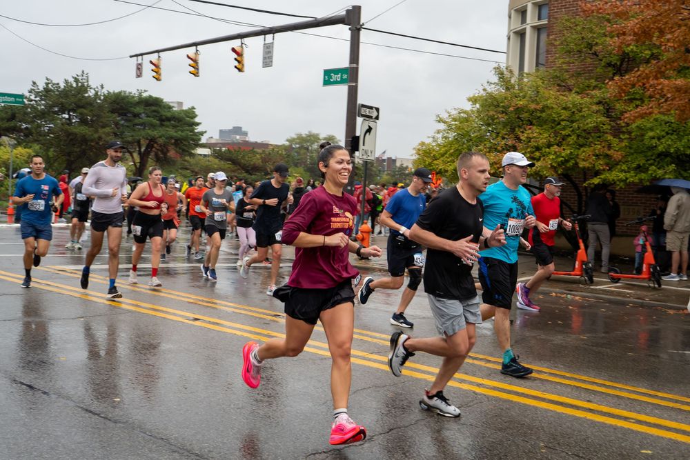 Runners participate in a rainy marathon on a wet city street lined with autumn trees and traffic lights.