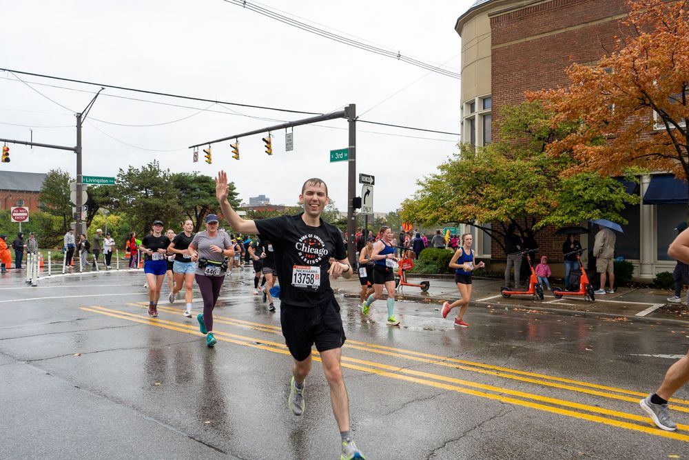 Runners compete in a rainy marathon race along a wet city street with fall foliage in the background.