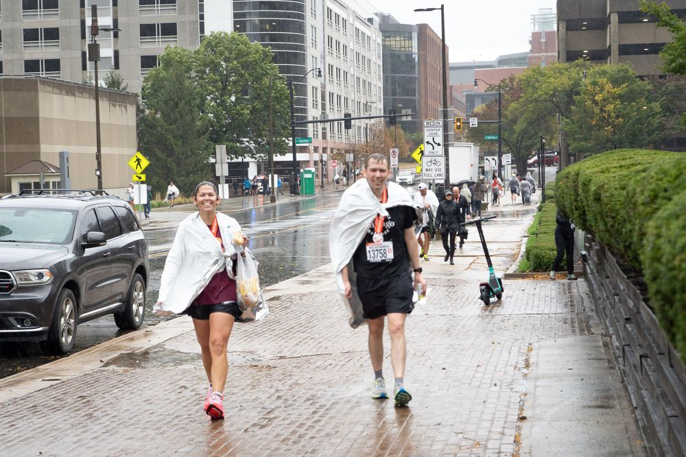 Runners jog along a wet city sidewalk on a rainy day in an urban setting with buildings and parked cars nearby.