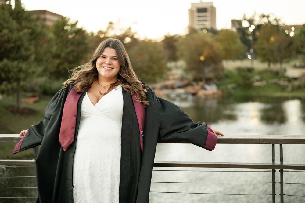 A person wearing a maroon jacket and white sweater smiles while standing on a bridge during golden hour.