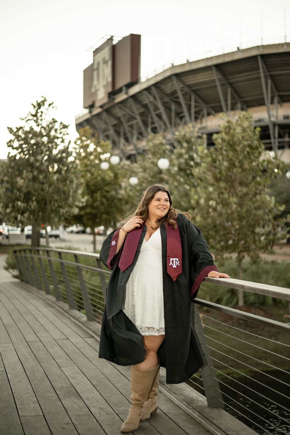 A graduate poses in front of a stadium wearing a black jacket and maroon scarf during a college photo session.