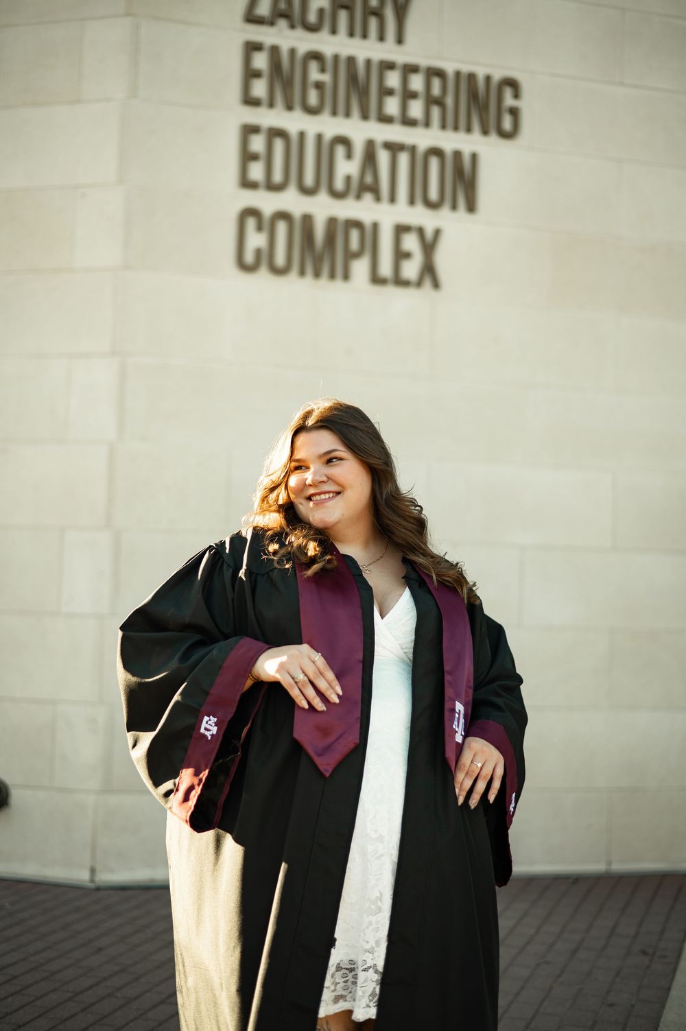 Graduate in black cap and gown poses joyfully against engineering building wall during graduation photoshoot.