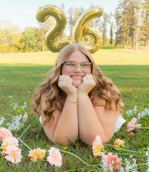 Birthday photo with gold number 26 balloons surrounded by pink and peach flowers on grass.