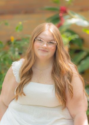 Young woman with long auburn hair and pink glasses smiling outdoors in white top with green foliage background.