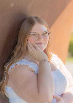 Young woman with glasses and long blonde hair poses against a rustic wall in a white top, smiling warmly.