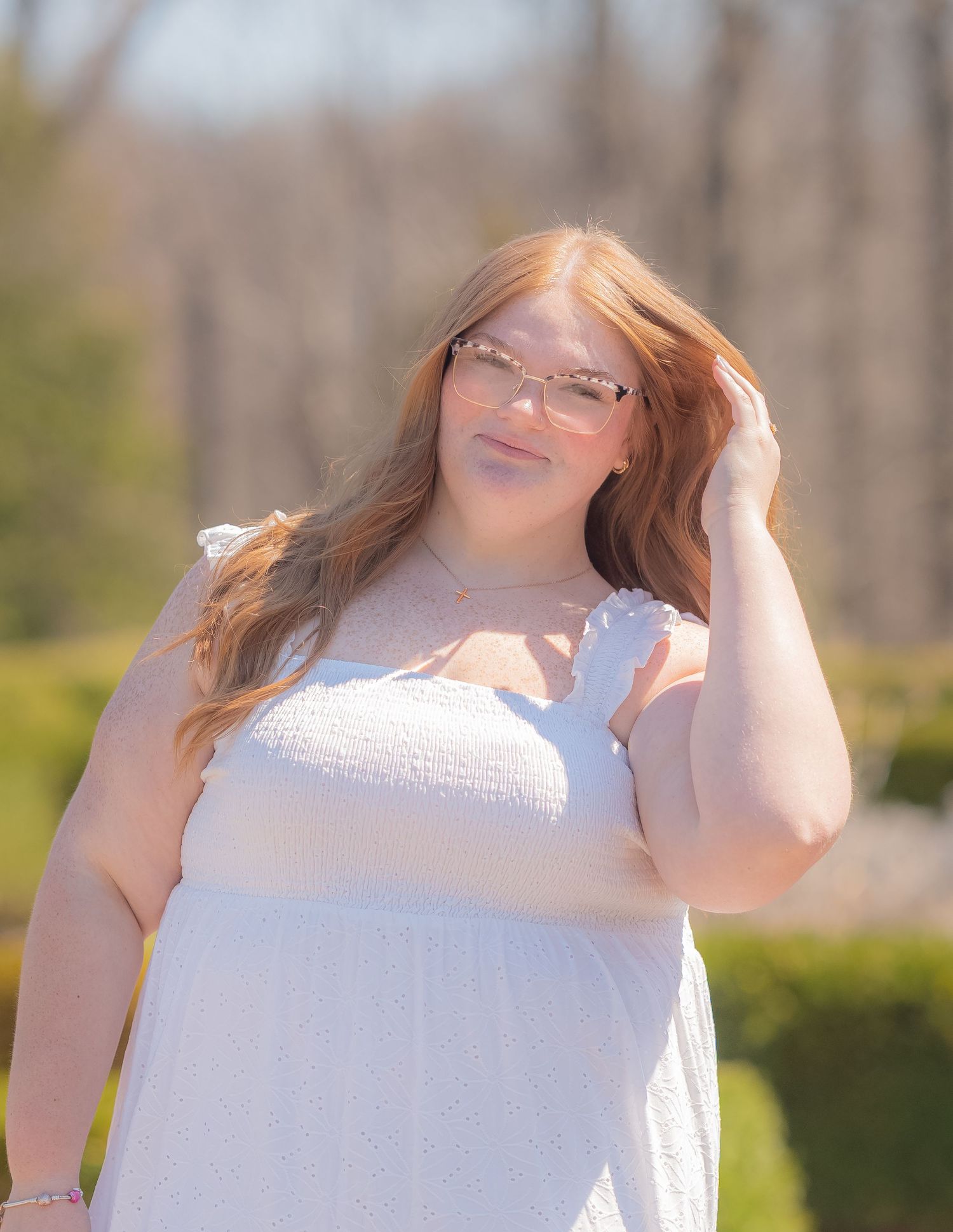Young woman with red hair in white dress and pink glasses poses outdoors in sunny garden setting.
