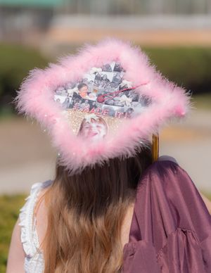 Decorated graduation cap with pink feather boa trim and collage of photos, worn by a graduate in a mauve gown.