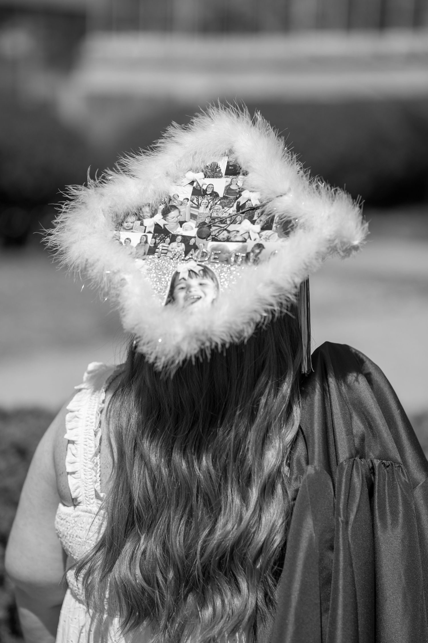 Graduate with decorated mortarboard cap adorned with feathers and photos, wearing gown, viewed from behind.