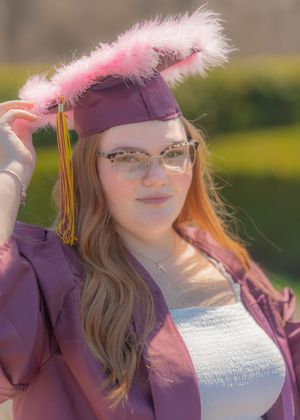 Young woman in maroon graduation cap with pink feathers and gown, wearing glasses, posing outdoors.