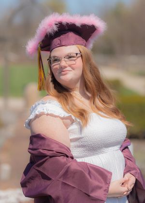 Young woman in maroon graduation cap with pink feathers and gown posing outdoors for graduation photos.