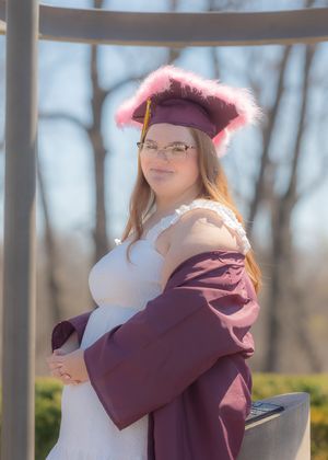 Graduate in maroon cap and gown with pink feather trim poses outdoors for senior portrait photo.
