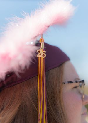 Close-up of a maroon graduation cap with pink feathers and a gold tassel charm reading 26.