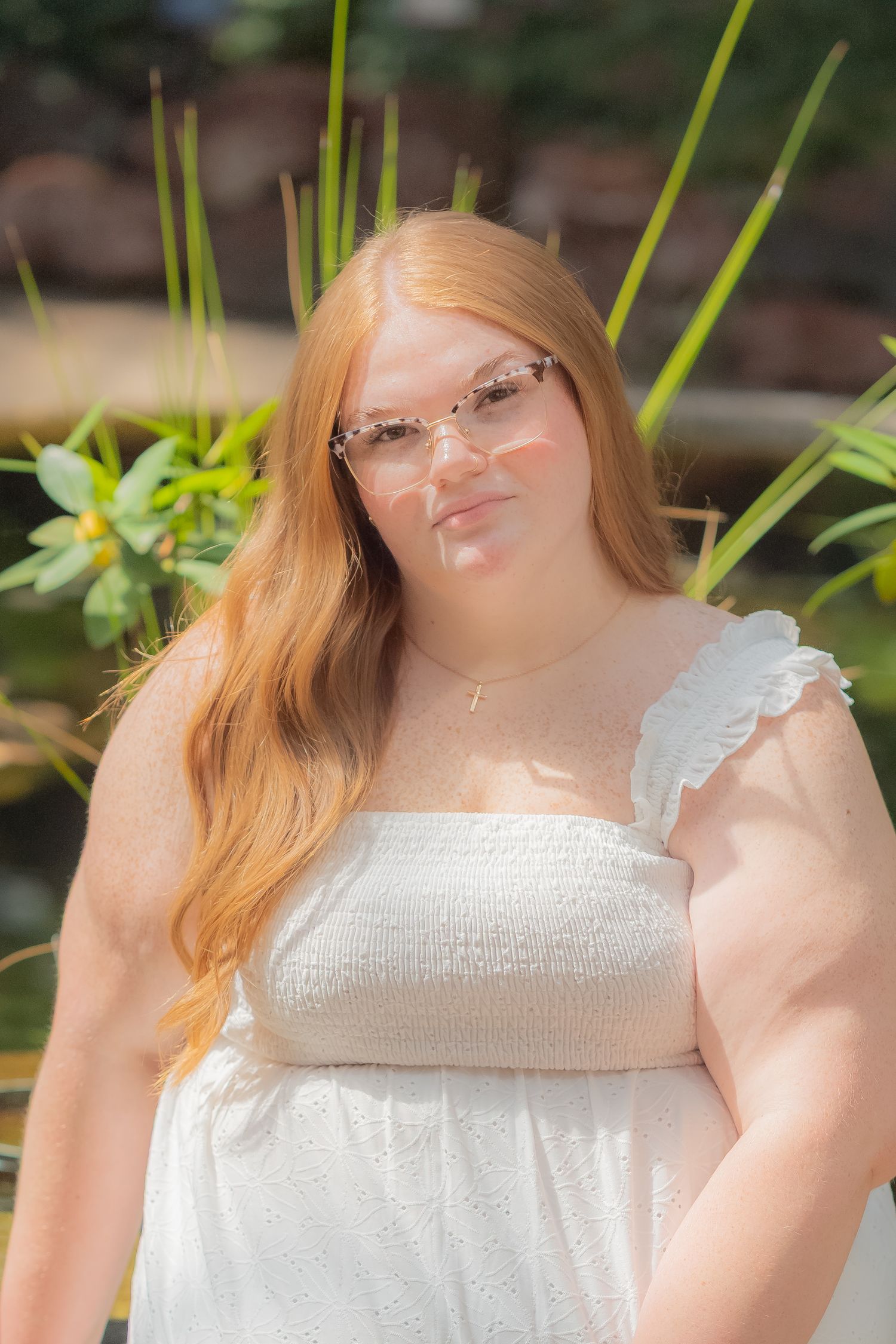 Young woman with long red hair wearing white dress and pink glasses posing outdoors near tropical plants.