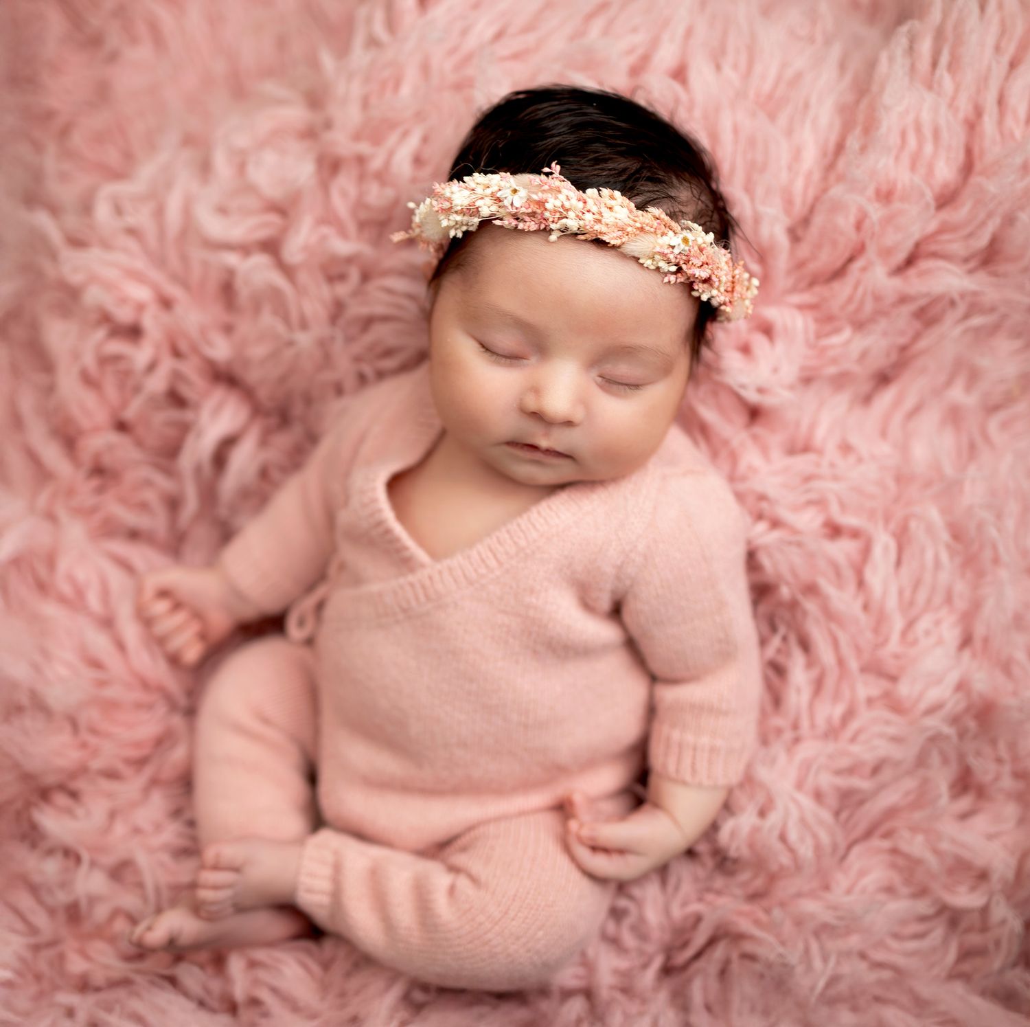 Newborn baby wearing pink knitted outfit sleeping peacefully on soft pink furry blanket.