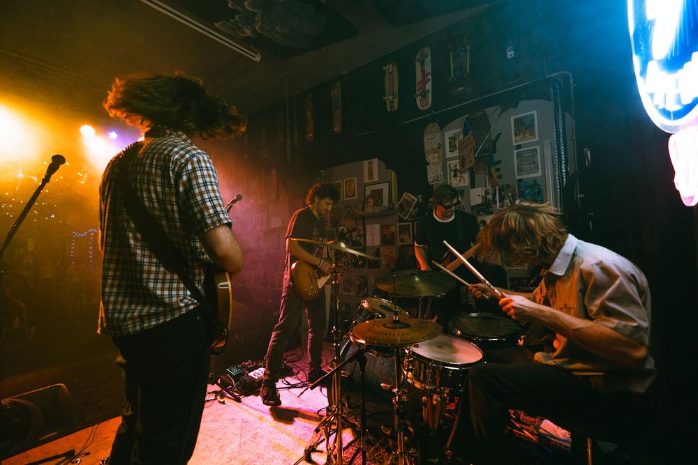 Band performing on stage in a dimly lit venue with guitarist, bassist, and drummer playing live music.