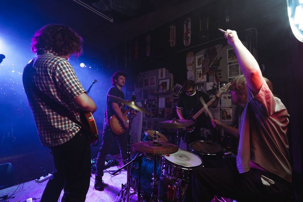 Band performs on a small club stage with blue lighting, drummer visible in the background surrounded by posters.