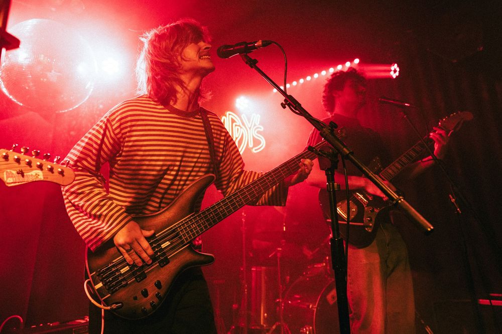 Two band members perform on stage with red concert lighting, one playing bass guitar in a striped shirt.