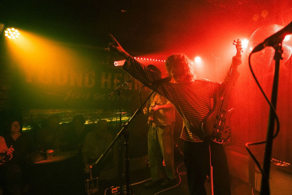 Band performs energetically on a dimly lit stage with red and yellow lights, one member raising their arm triumphantly.
