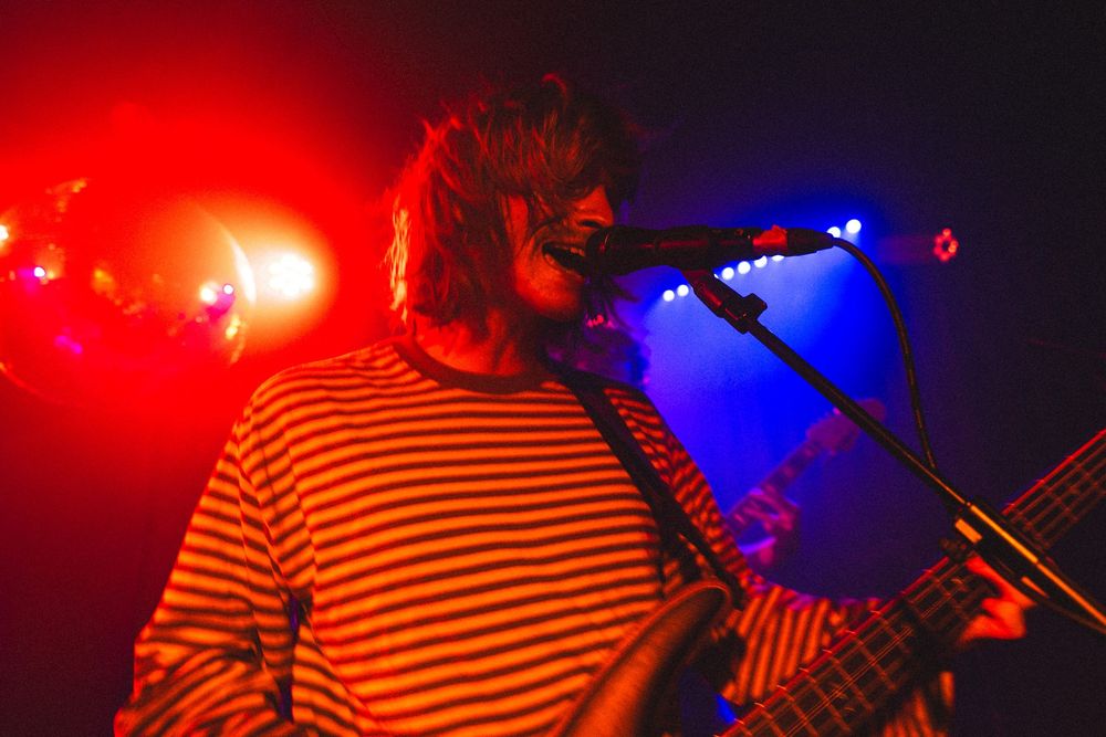 Male vocalist in a red and white striped shirt sings passionately into a microphone under vivid red stage lighting.