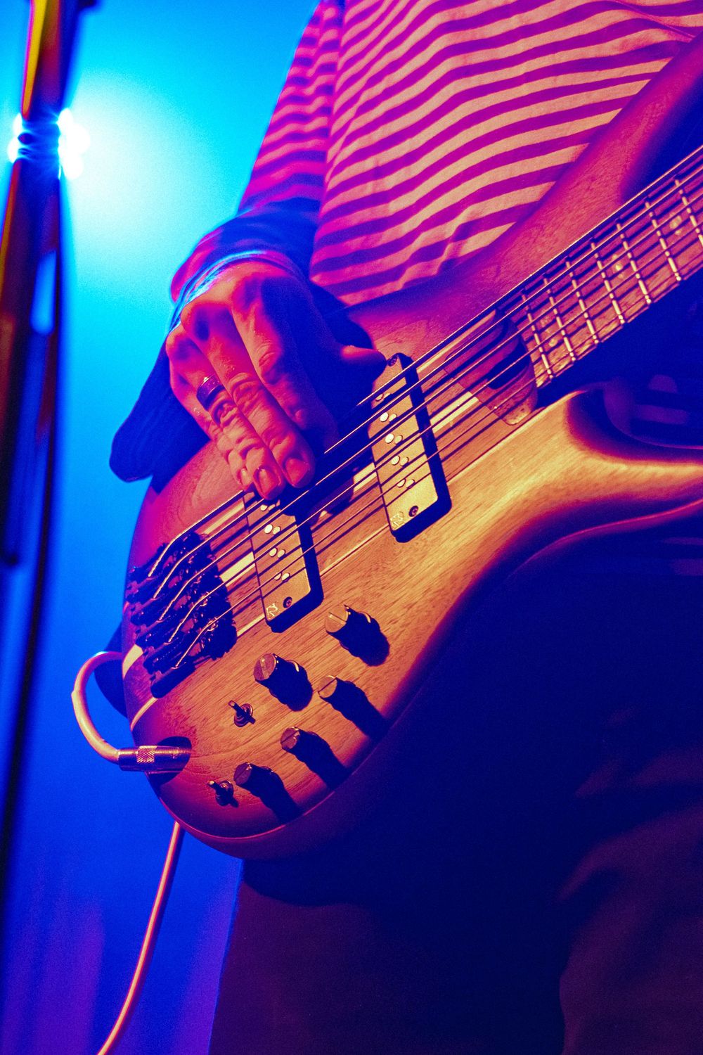 Close-up of a musician's hand playing a brown bass guitar under blue and red stage lighting at a live show.