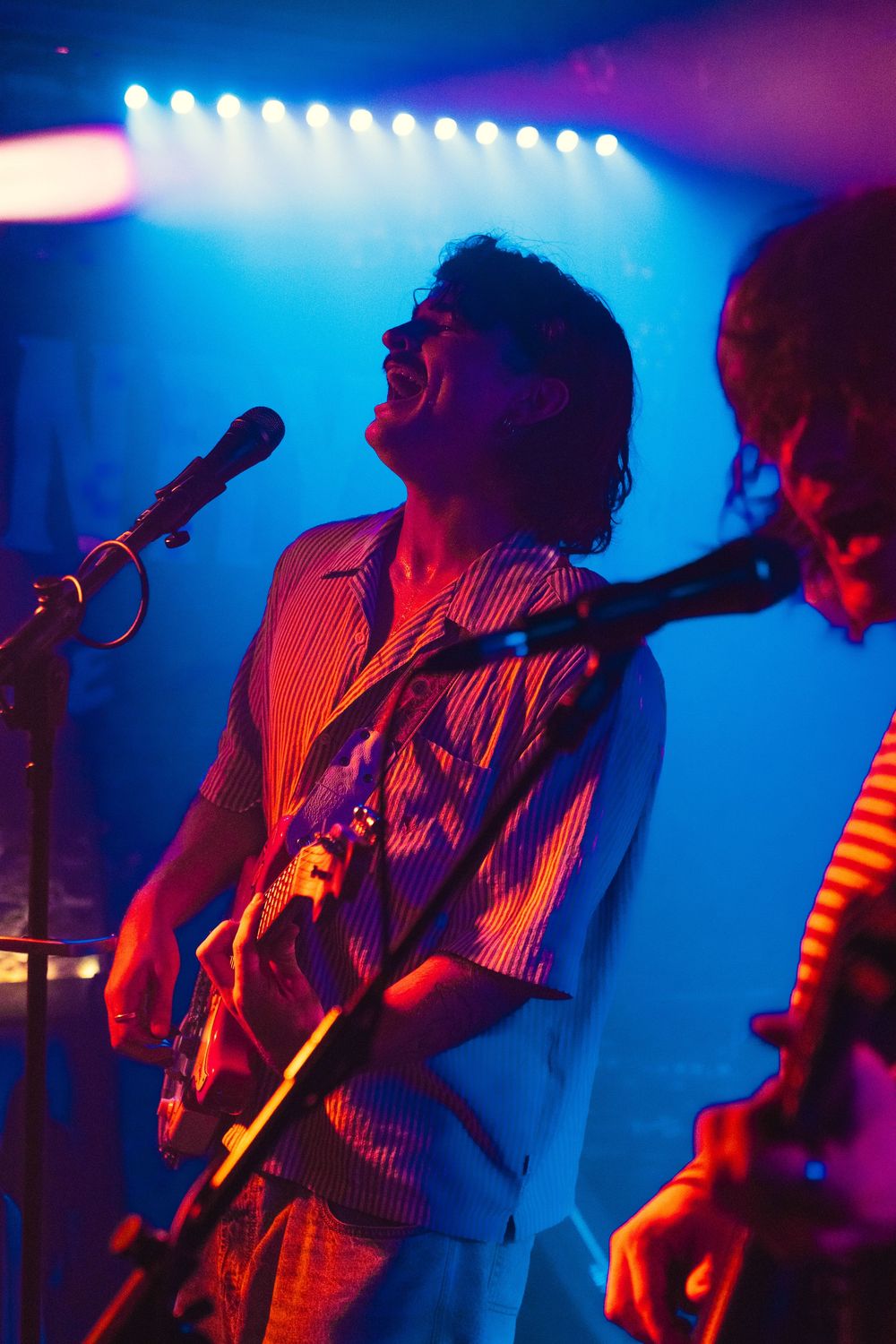 Male guitarist sings into a microphone while playing electric guitar under blue and purple concert stage lighting.