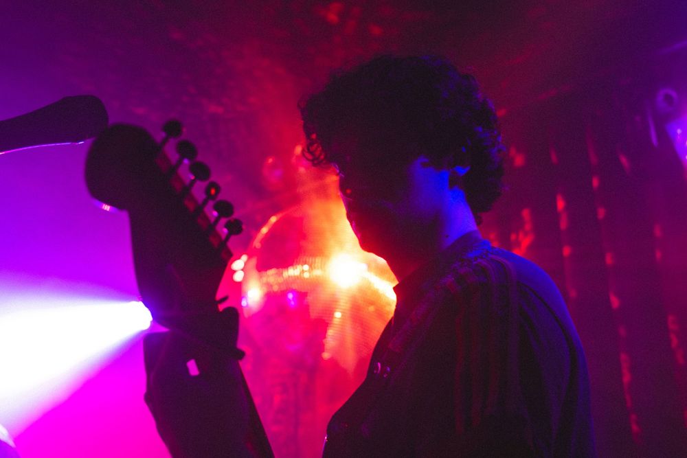 Silhouetted musician holds a guitar against vivid purple and red backlit stage lighting at a live performance.