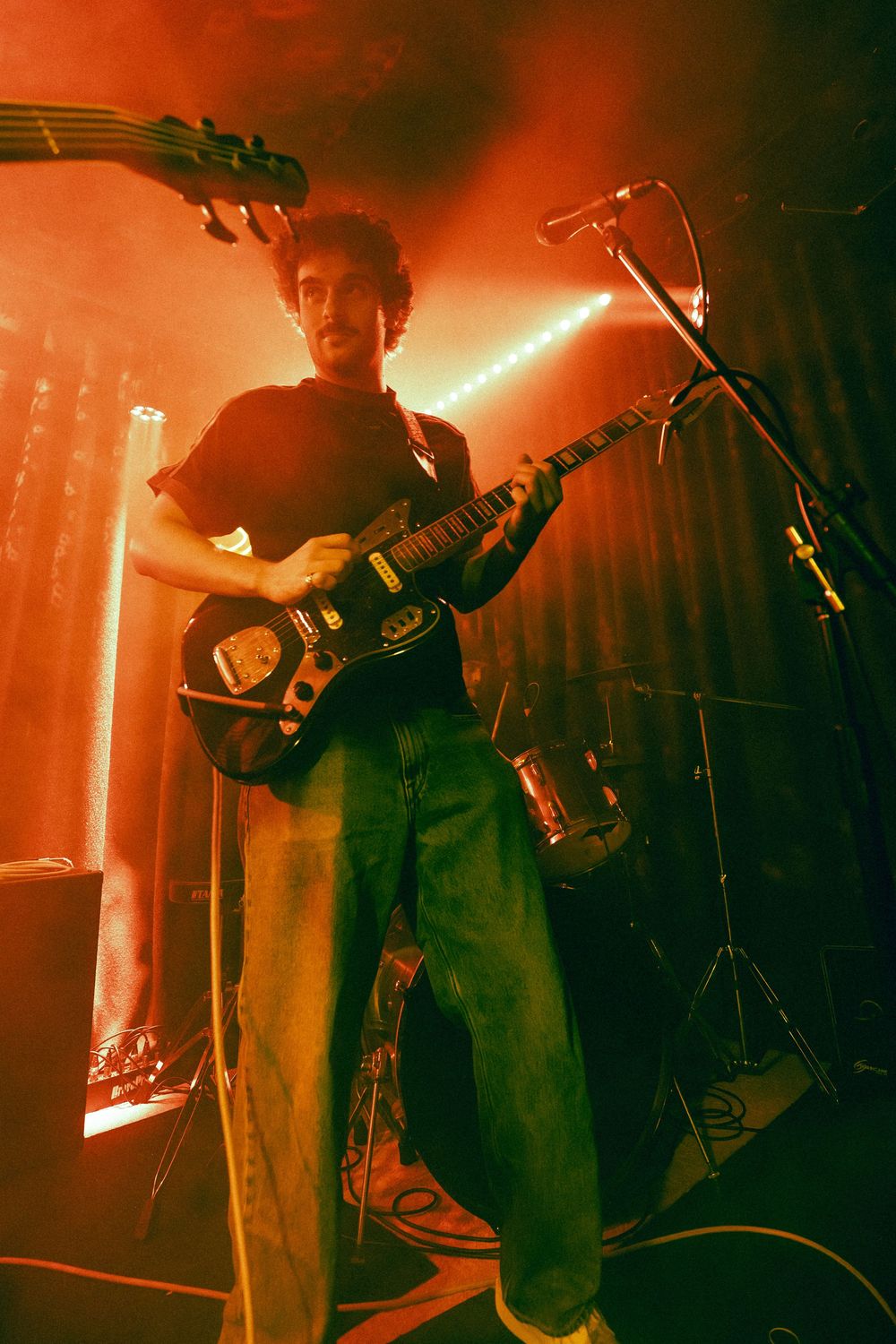 Young male guitarist plays electric guitar on stage bathed in warm red atmospheric lighting at a live music venue.