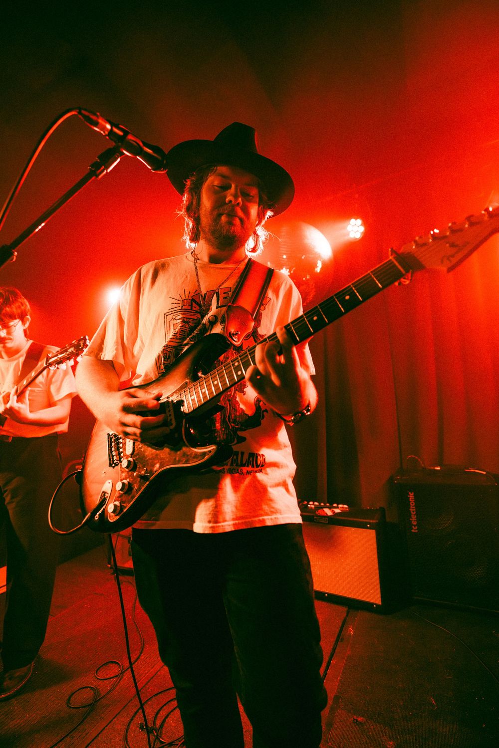 Guitarist in wide-brim hat plays electric guitar on stage under dramatic red lighting at live concert.