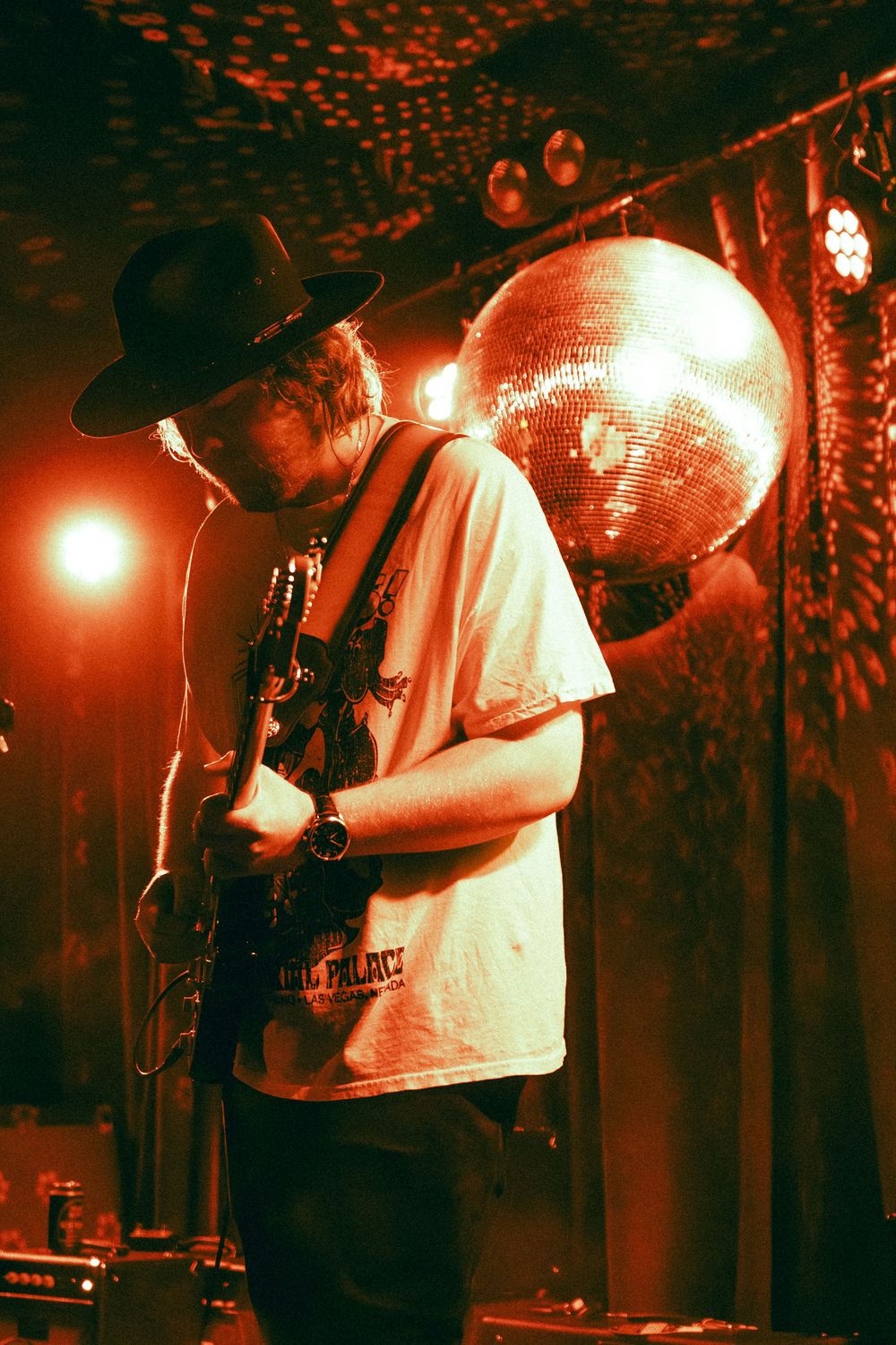 Male guitarist in black hat plays guitar on stage bathed in red light with disco ball behind him.
