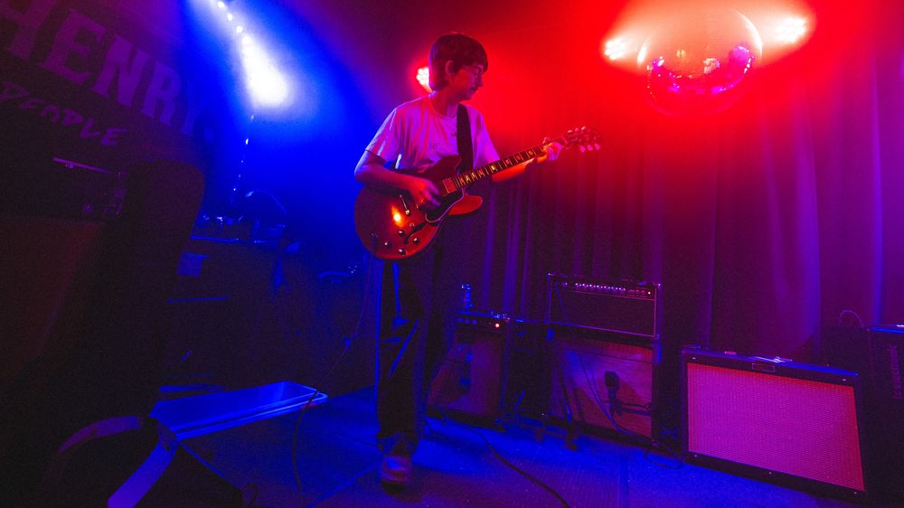 Solo guitarist performs on stage under vibrant blue and red stage lights in a small music venue.